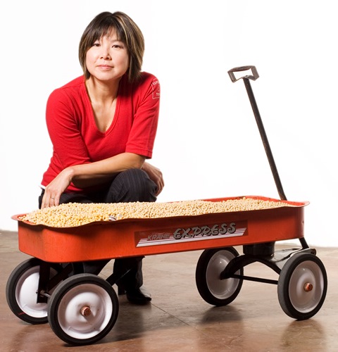 A confident asian woman in a red top kneeling by a red kids wagon full of soybeans